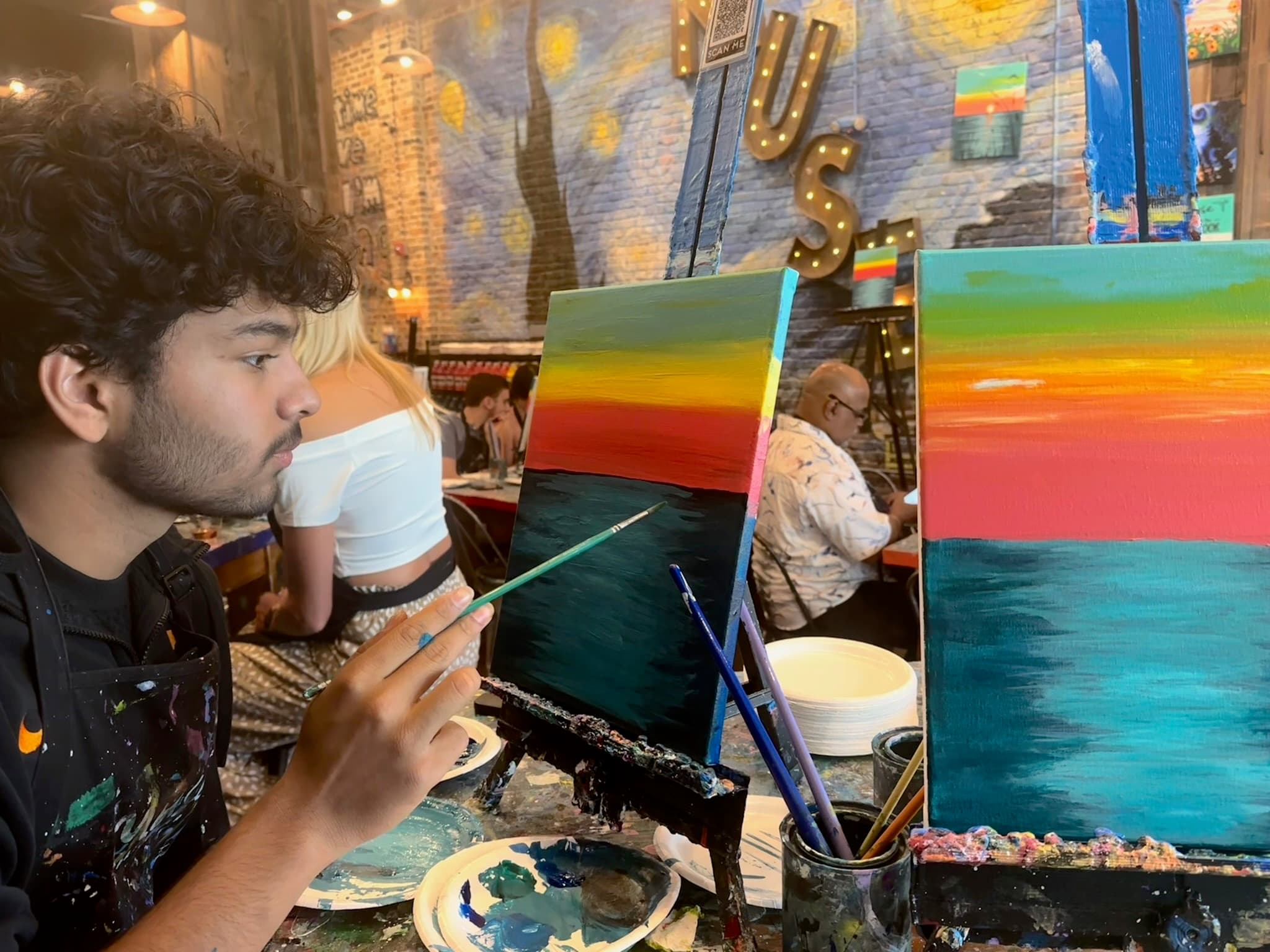 A painter in a paint-spattered apron working on a canvas of a sunset over water, a finished version of the same scene on the easel beside it