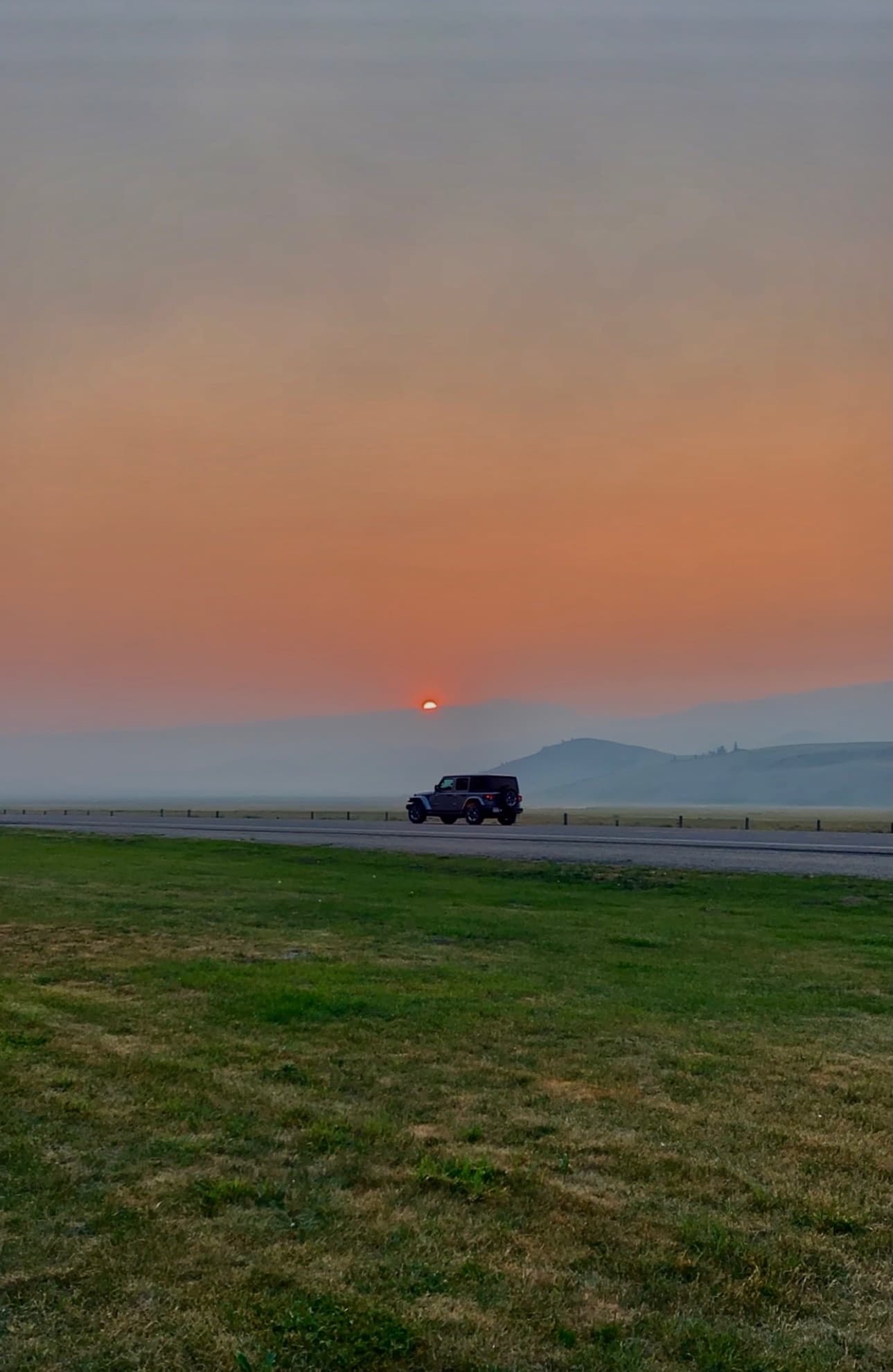 A Jeep on an empty highway across grassland, the sun a dim orange disc in a hazy sky